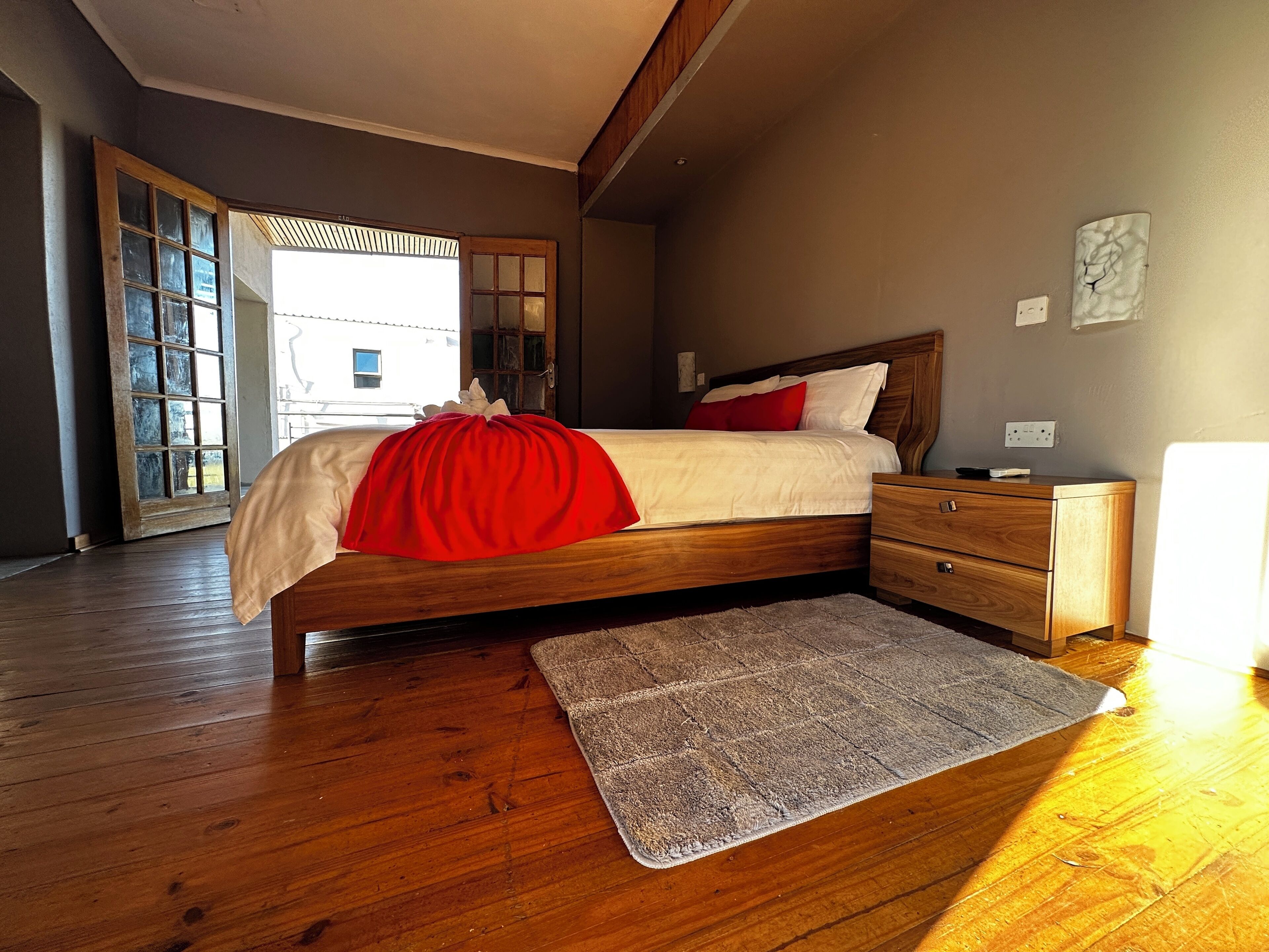 A bedroom with a wooden bed, a nightstand, a bedside table, a rug, and a view of the outside.