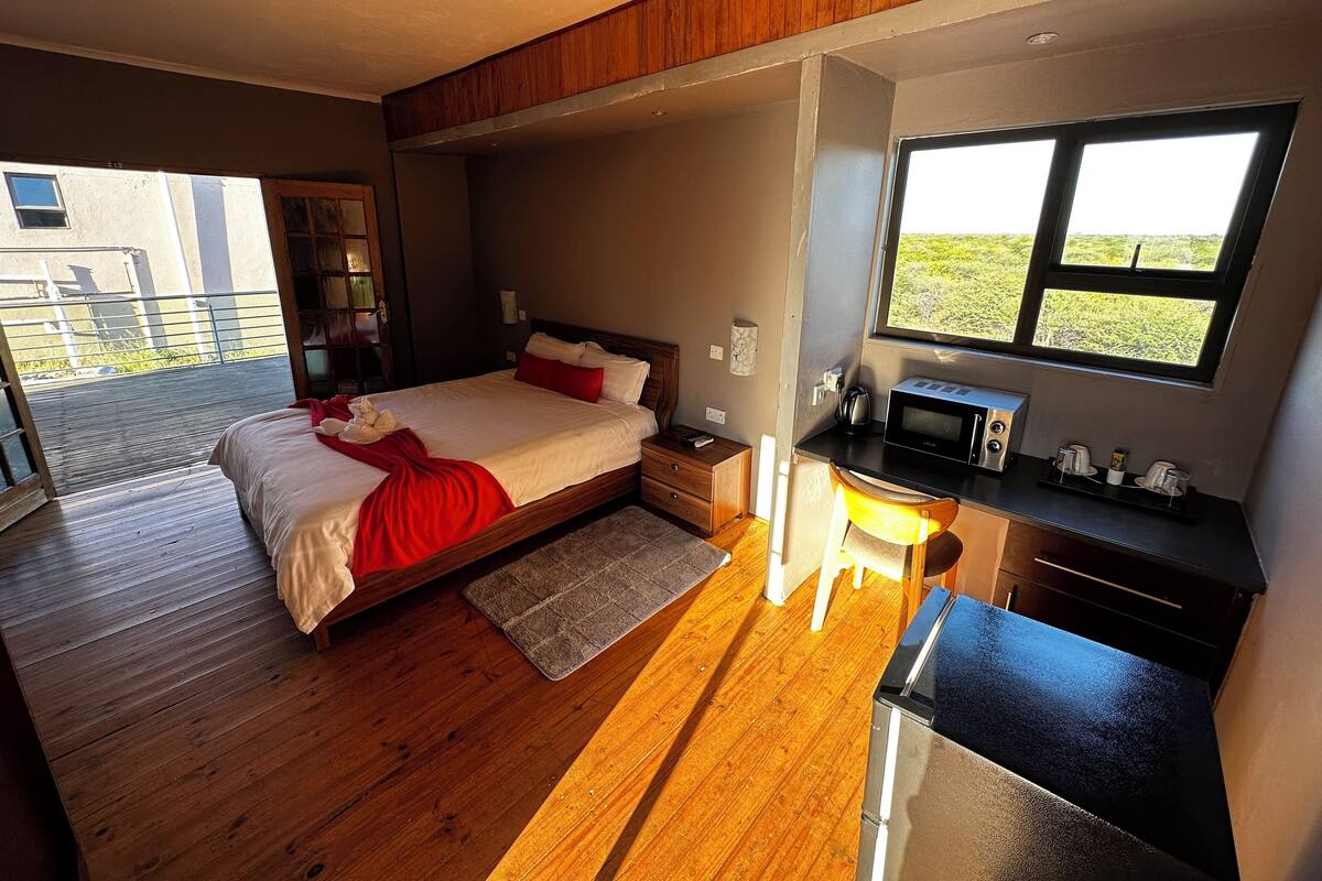 A modern bedroom with a wooden floor, a large bed with red bedding, a desk with a microwave, and a view of greenery outside.