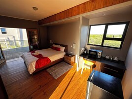 Un dormitorio moderno con piso de madera, una cama grande con ropa de cama roja, un escritorio con microondas y vistas a zonas verdes.