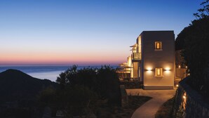 A modern building with a balcony overlooking the sea, a pathway leading to the entrance, and a view of the ocean at dusk. - Odyssey Rock (Milos)