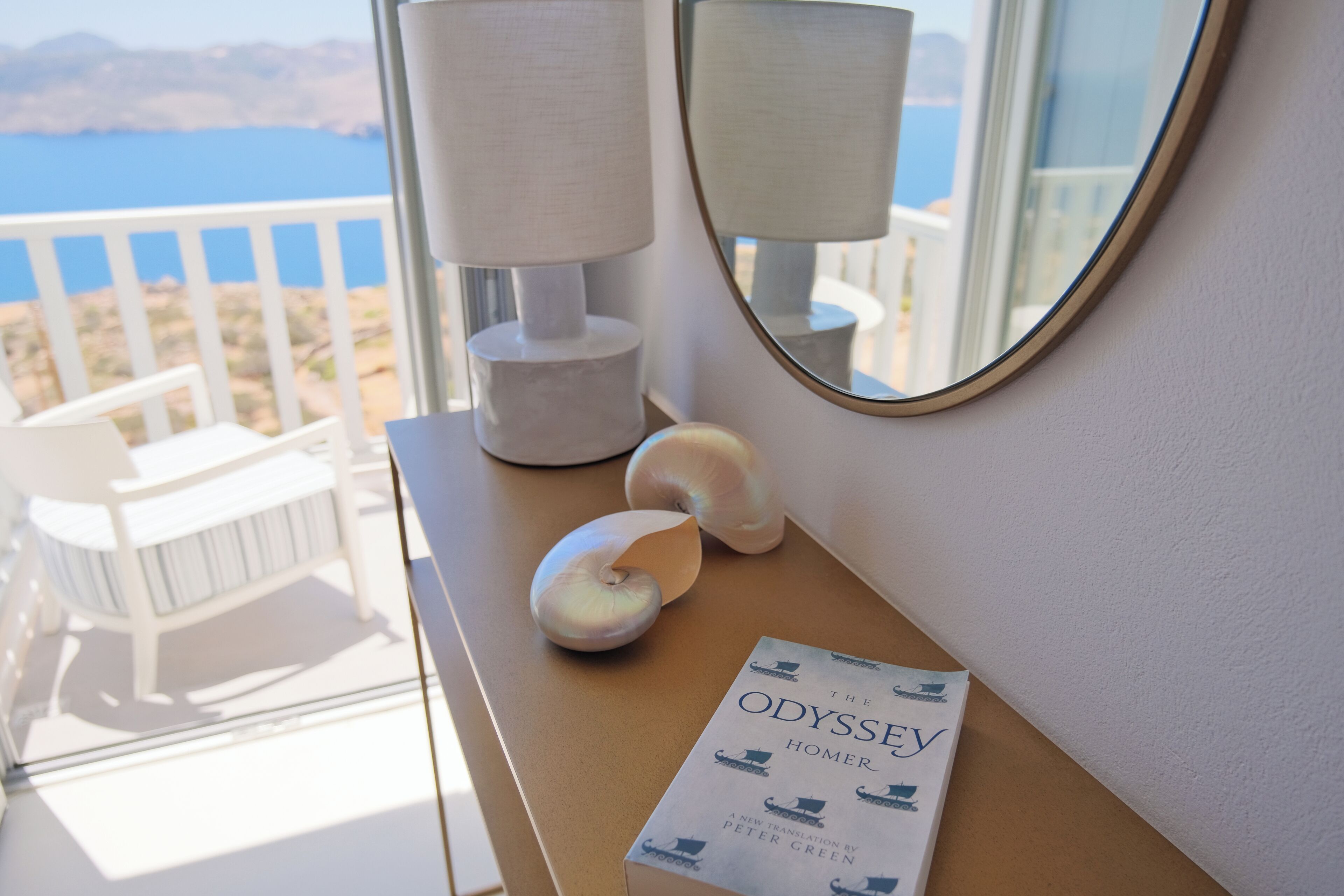 Una habitación con vista al mar, un libro titulado 'La Odisea' de Homero, una lámpara y un espejo.