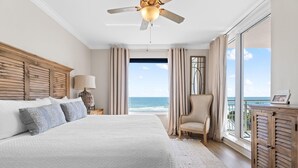 A bedroom with a large bed, a ceiling fan, a nightstand with a lamp, a chair, and a view of the ocean. - Indigo East 406 (Pensacola)
