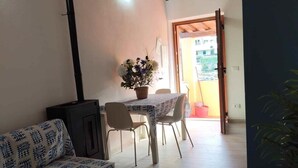 Un salon chaleureux avec un canapé, une table entourée de chaises, une cheminée et une vue sur l’extérieur par une porte ouverte.