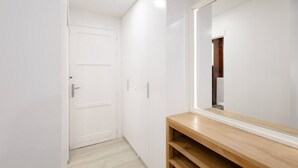 Un couloir étroit avec une porte blanche et un grand miroir qui reflète une étagère en bois.