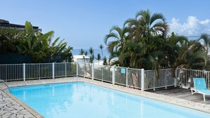 Une piscine entourée d’une clôture blanche, bordée de palmiers et surplombée d’un ciel dégagé.