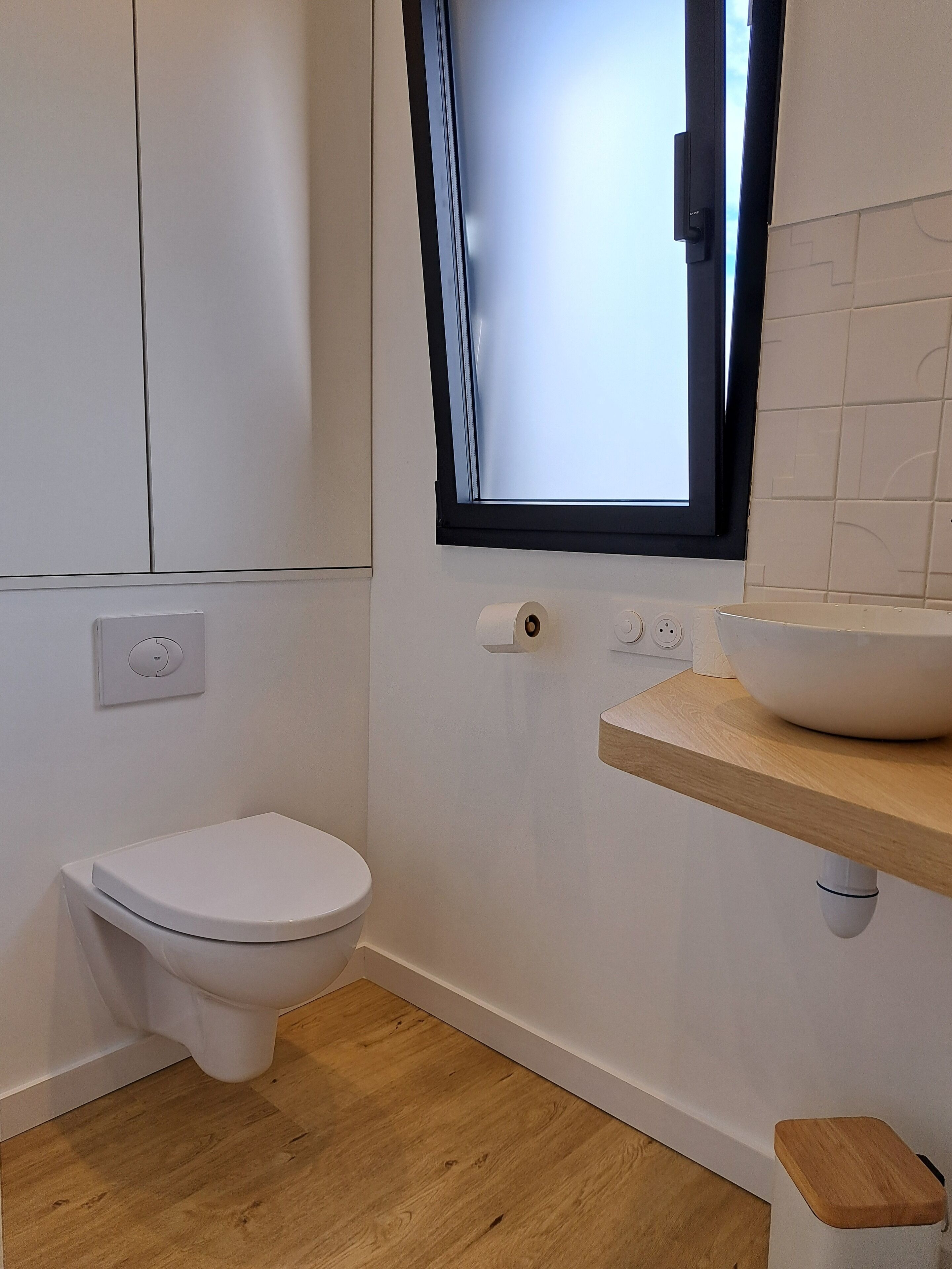 Une salle de bain moderne avec des toilettes blanches, un meuble-lavabo en bois doté d’un vasque, une fenêtre à cadre noir et un plancher en bois.