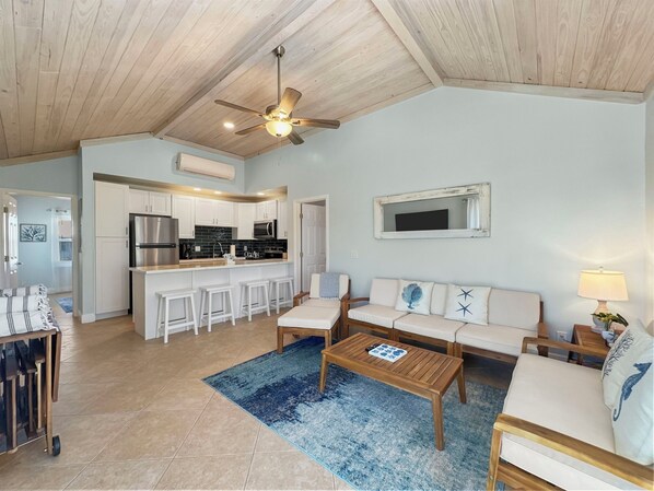 Uma sala de estar ampla com teto de madeira, ventilador de teto, cozinha com armários brancos, área de bar e espaço de estar com tapete azul.