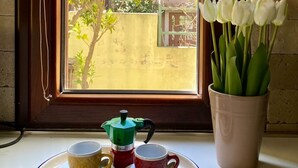 Una ventana con vistas a un árbol y un edificio, una persiana, un picaporte y una mesa con una cafetera y un jarrón de flores blancas.