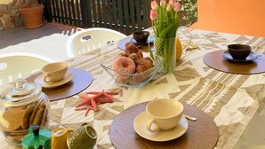 Un área de comedor al aire libre con una mesa preparada para dos personas, que incluye una variedad de pasteles, una cafetera y un florero con flores.