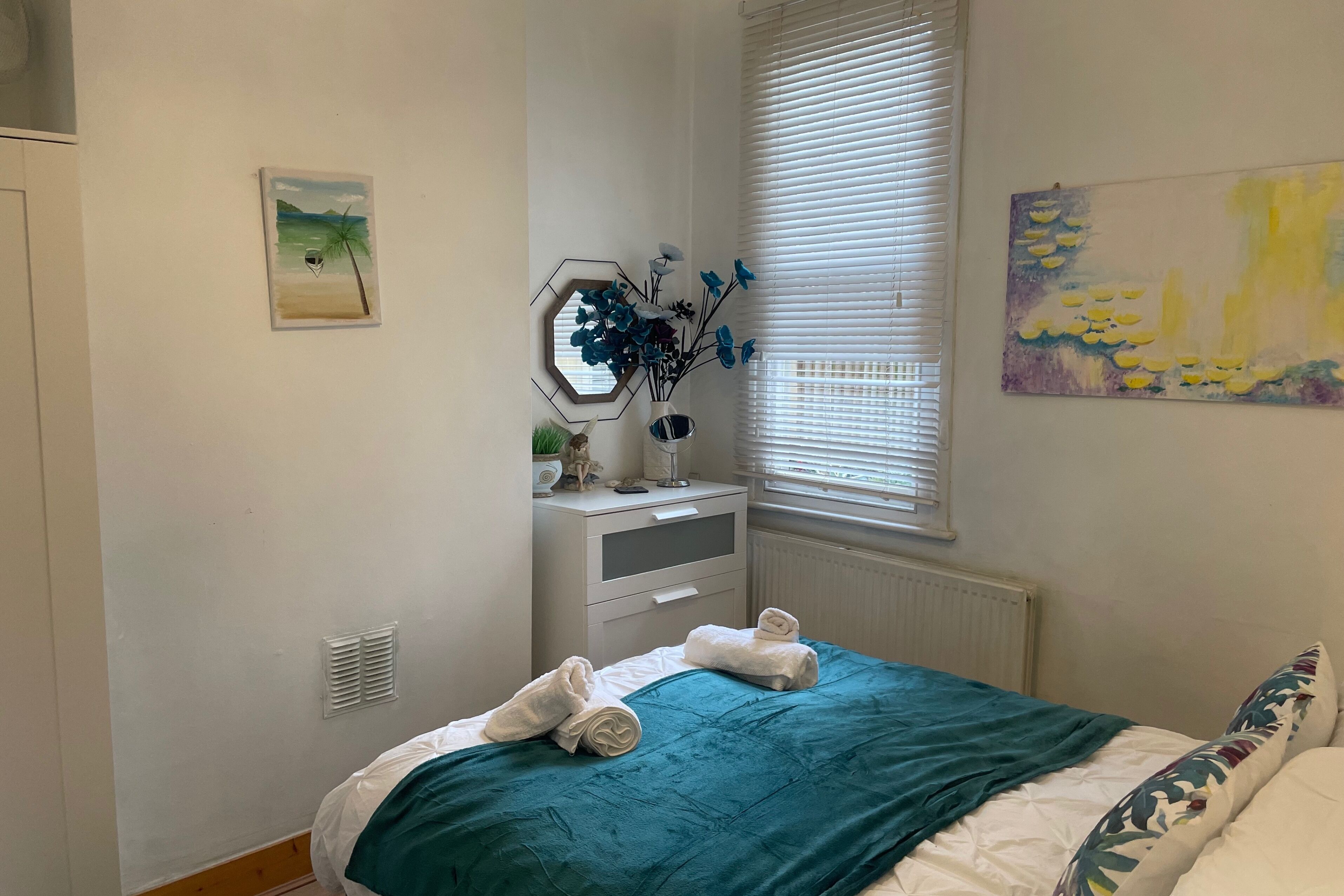 A bedroom with a bed, a nightstand, a window with blinds, and wall art.