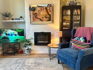 A living room with a flat-screen TV, fireplace, bookshelf, and a blue armchair. - 1 Bed Victorian Flat Sleeps 4 - Garden & Patio (London)