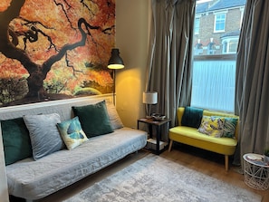 A living room with a sofa, a small table with a lamp, a window with curtains, and a wall with a tree mural.