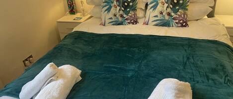 A neatly made bed with a teal bedspread, white pillows, and a floral-patterned pillow. There are two white towels folded at the foot of the bed.