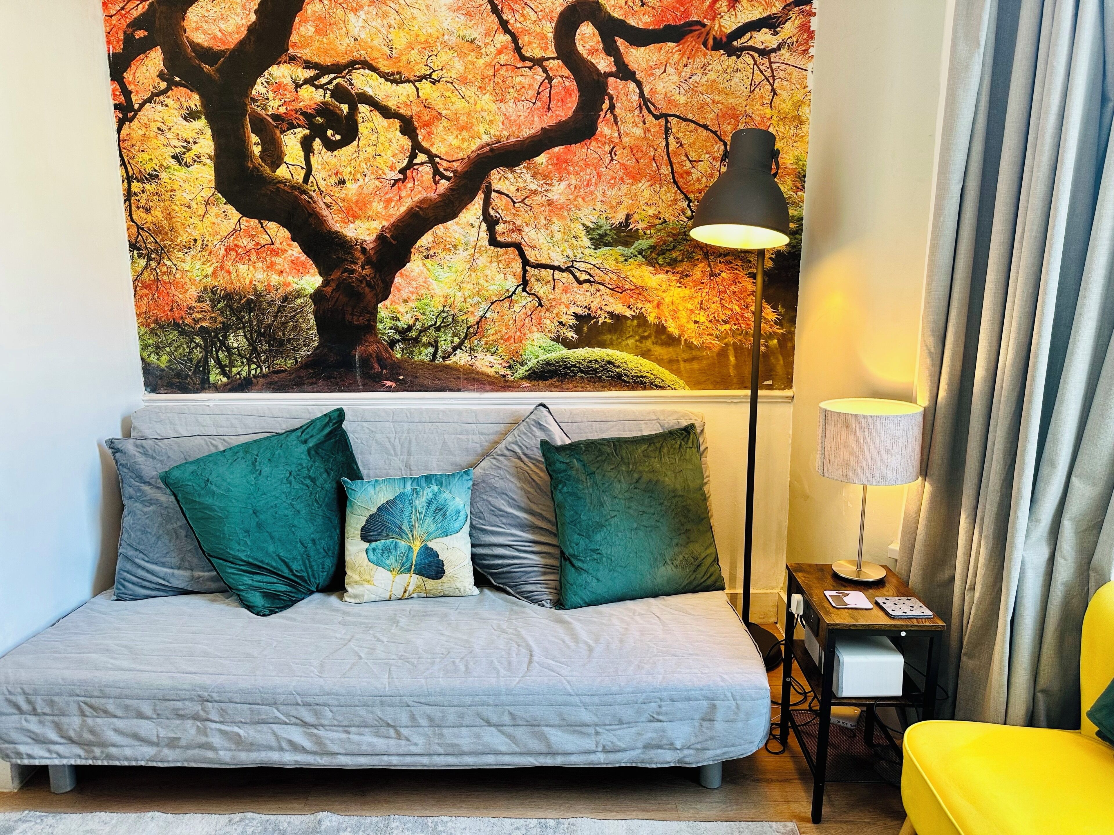 A bedroom with a bed, pillows, a lamp, a desk with a lamp, and a large wall mural of a tree.