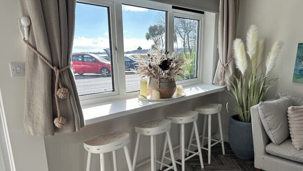 Un coin cuisine avec une fenêtre donnant sur un parking, un plan de travail blanc avec des tabourets et une plante en pot.