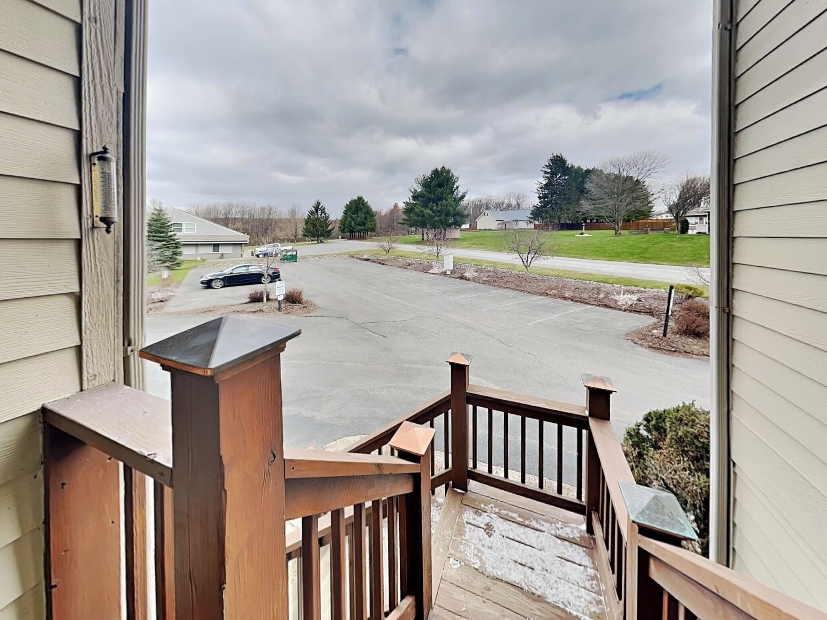 A view from a wooden deck to a parking lot with a car and a truck, surrounded by trees and a cloudy sky.