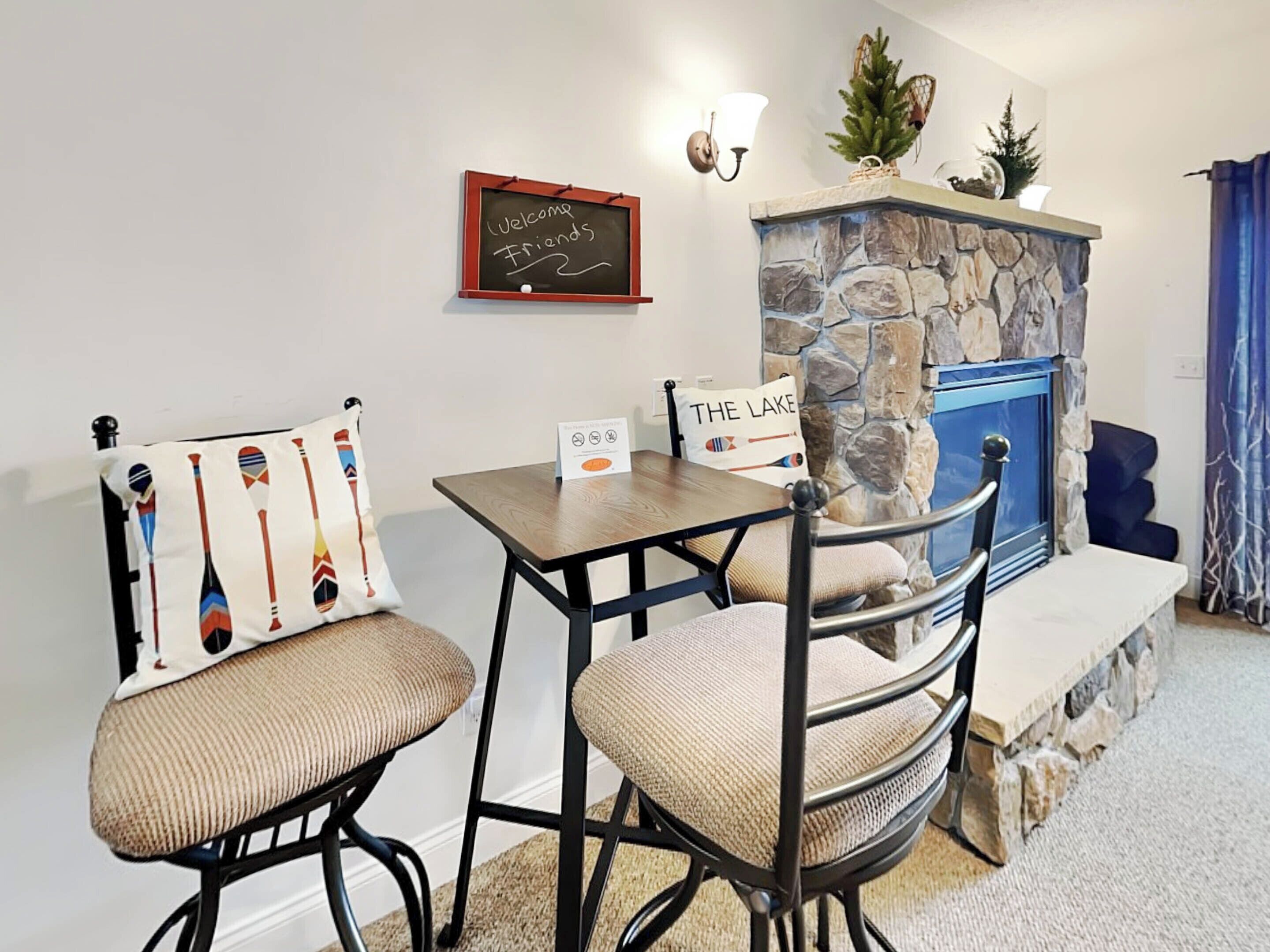 A cozy room with a stone fireplace, two chairs with cushions, a small table, and a welcome sign on the wall.