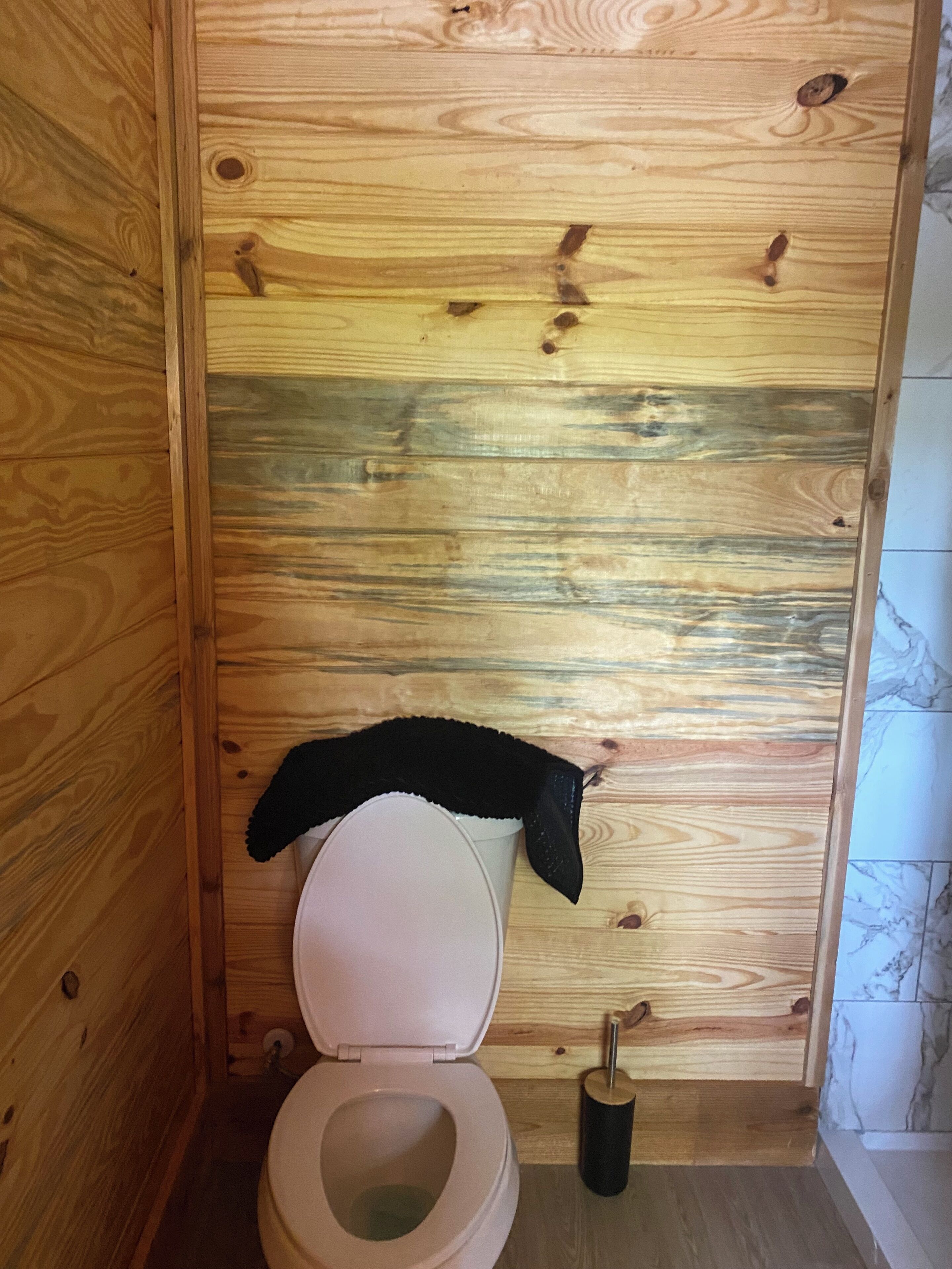 Een betegelde badkamer met een toilet, een zwarte doek over de toiletbril en een toiletborstel.