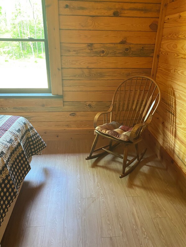 A wooden cabin room with a rocking chair, a bed with a patterned bedspread, and a window with a view of trees. - Cabin on Gaston. Fishing, hunting, relaxation in luxury. (Boydton)