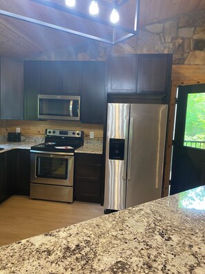 A kitchen with dark wooden cabinets, stainless steel appliances including a microwave and refrigerator, granite countertops, and a stone wall. - Cabin on Gaston. Fishing, hunting, relaxation in luxury. (Boydton)