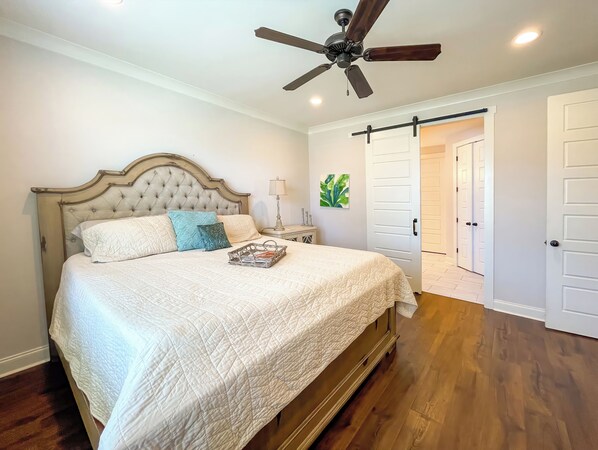 A bedroom with a bed, a ceiling fan, a nightstand, and a door leading to another room.