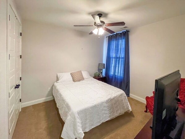 A bedroom with a bed, a ceiling fan, a nightstand with a lamp, a TV, and a window with blue curtains. - Spacious house in a quiet community in Oxford, Ms. Pet Friendly. (Oxford)