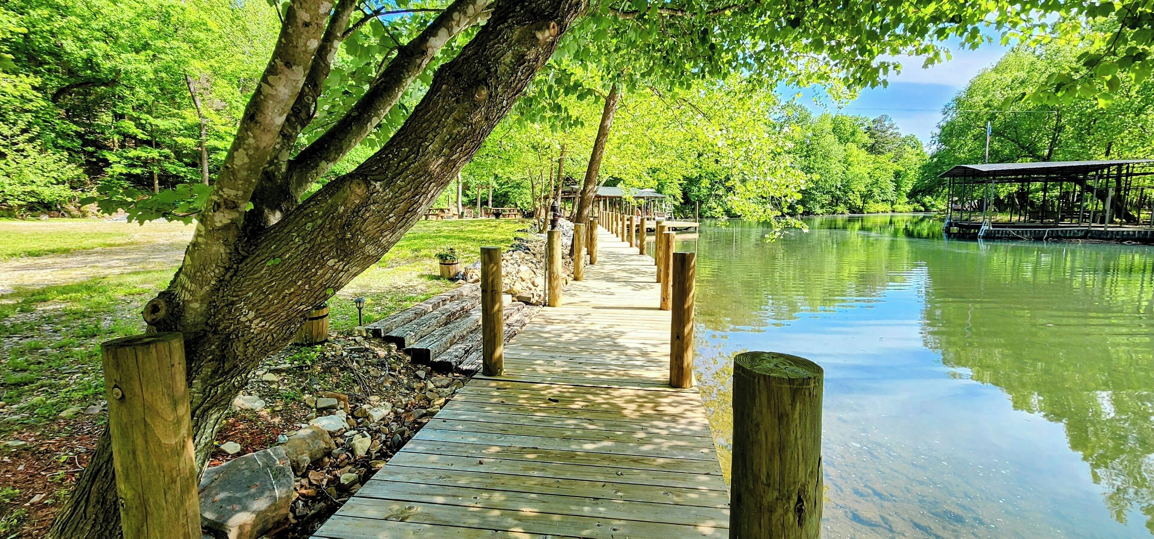 A wooden boardwalk leading to a covered pavilion by a calm lake, surrounded by lush greenery.