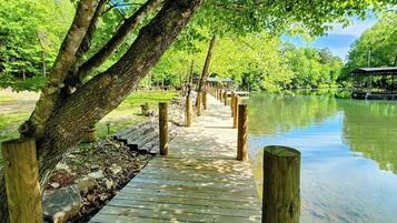 A wooden boardwalk leading to a covered pavilion by a calm lake, surrounded by lush greenery.
