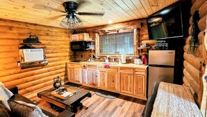 Ein gemütliches Blockhauszimmer mit Holzwänden, einem Holzesstisch, einer Couch, einer Küchenzeile mit Holzausstattung und einem Kühlschrank, sowie einem Deckenventilator.