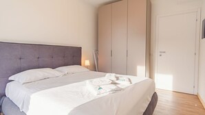 Een net opgemaakt bed met witte lakens en kussens, een grijs gestoffeerd hoofdbord en een grote inloopkast met schuifdeuren.