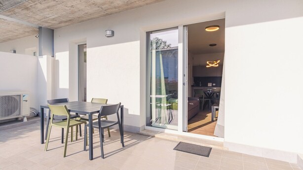 Een modern overdekt terras met een tafel en stoelen, een schuifpui naar een keuken en een plafondventilator.