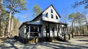 Une cabane dotée d’une grande véranda, de nombreuses fenêtres et d’un toit à forte pente, entourée d’arbres.