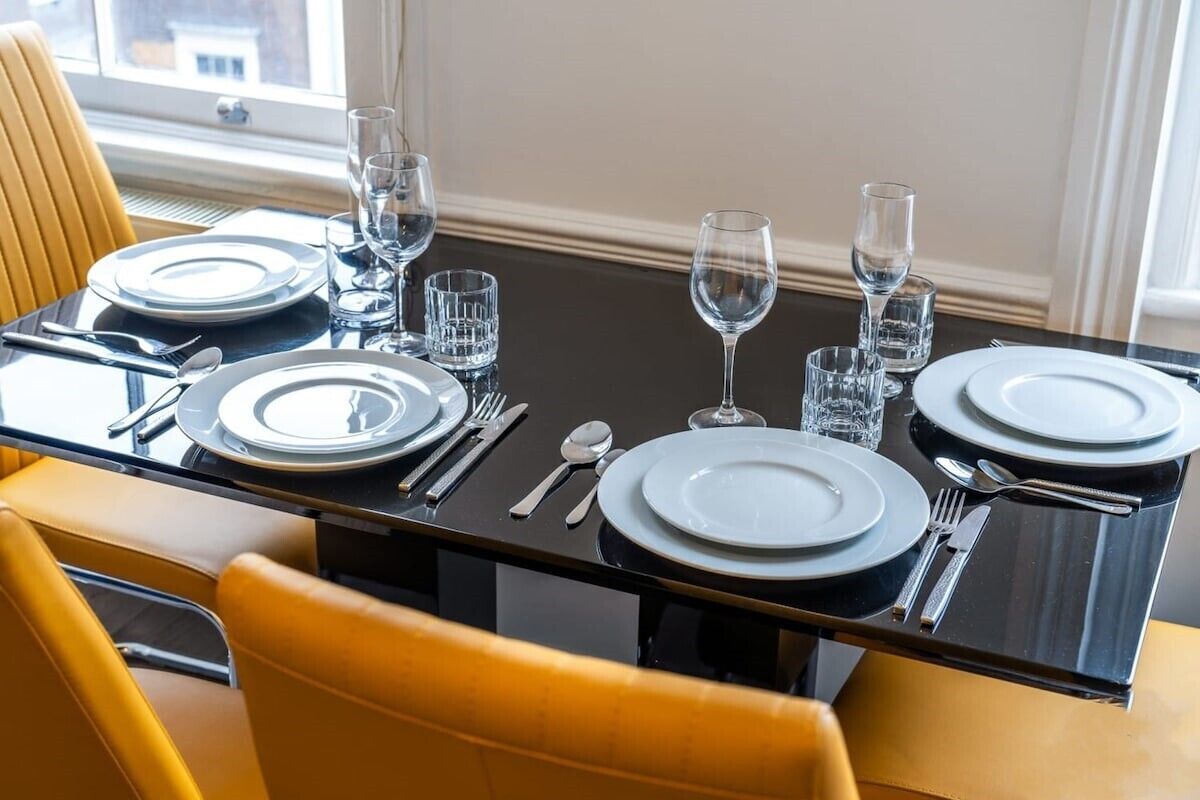 Una mesa de comedor dispuesta con platos, cubiertos y vasos, rodeada de sillas amarillas.