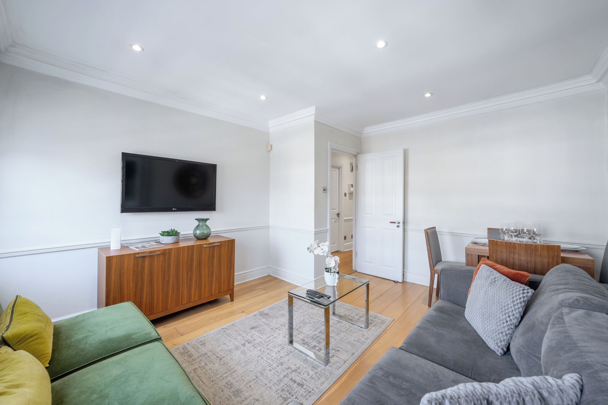 A modern living room with a flat-screen TV, wooden cabinet, green and grey sofas, a glass coffee table, and a dining area with a table and chairs.