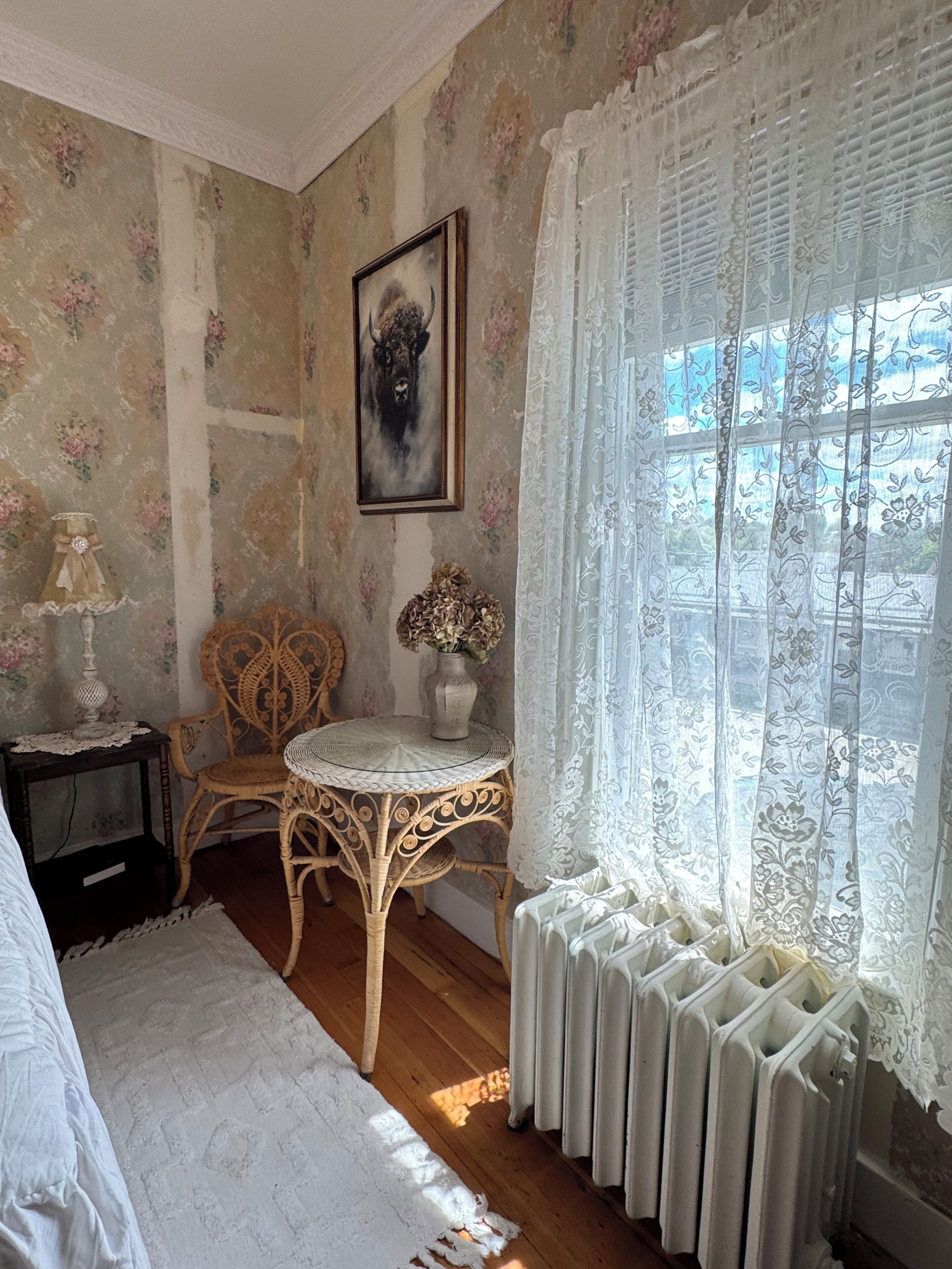 A room with floral wallpaper, a framed artwork, a wicker chair, a small table with a vase, and a radiator near a window with lace curtains.