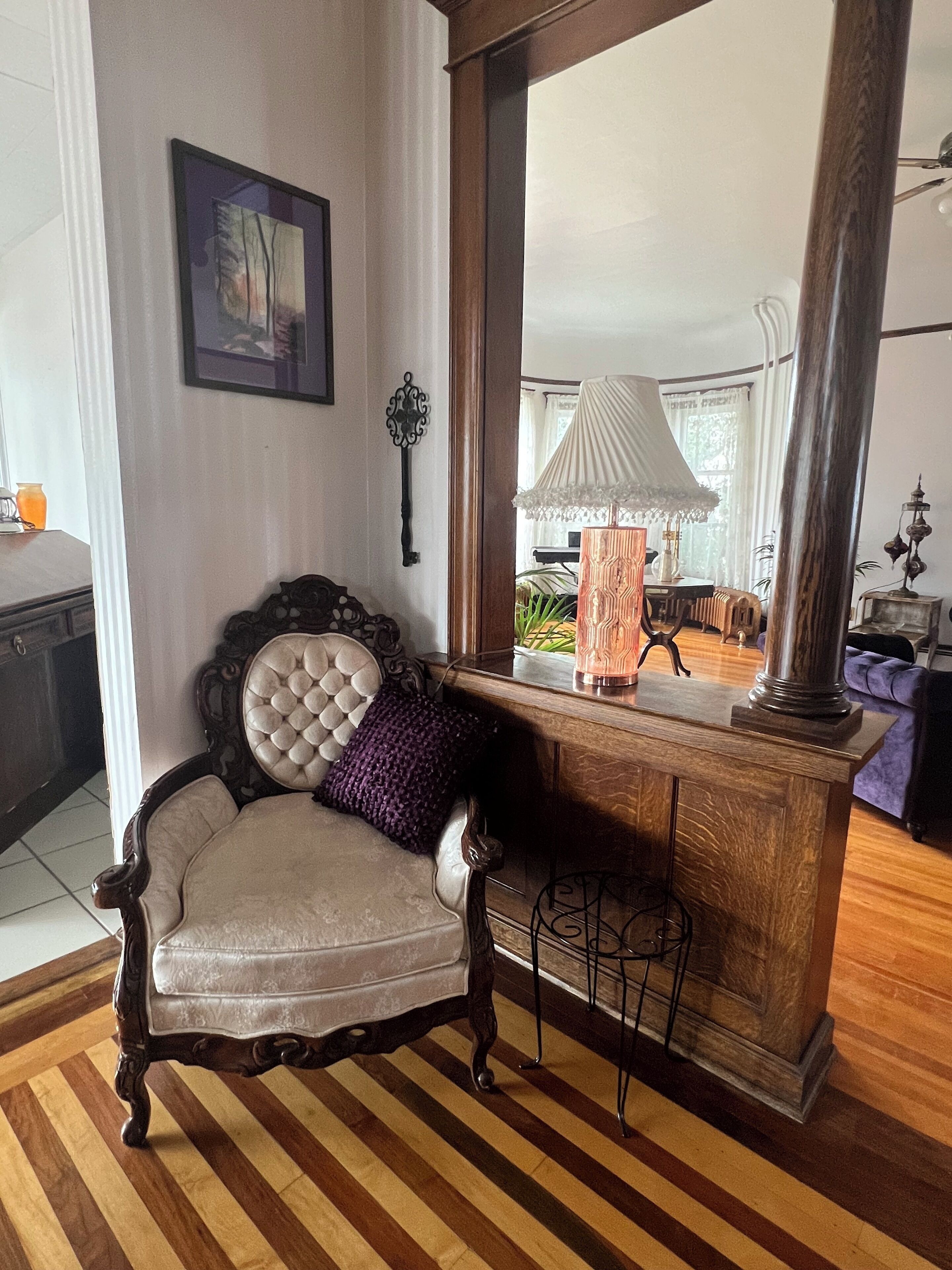 Una habitación con una silla de madera, una mesa con lámpara y un espejo grande que refleja la estancia.