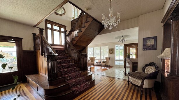 A grand staircase with a wooden finish and a chandelier, leading to a spacious living area with a patterned carpet and a large window. - The Boho Buffalo Inn (Buffalo)
