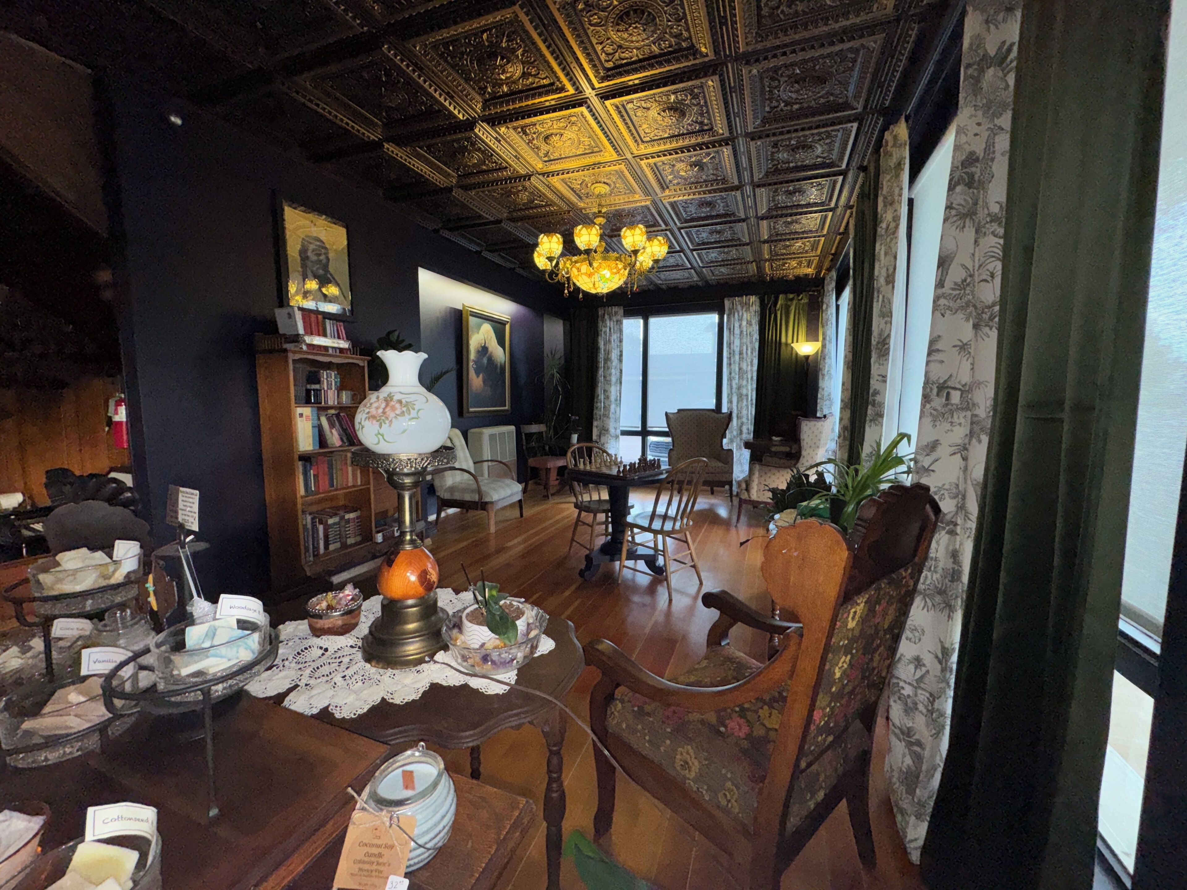 A room with a high ceiling, ornate ceiling design, a chandelier, a fireplace, a bookshelf, a dining table with chairs, and a large window with curtains.