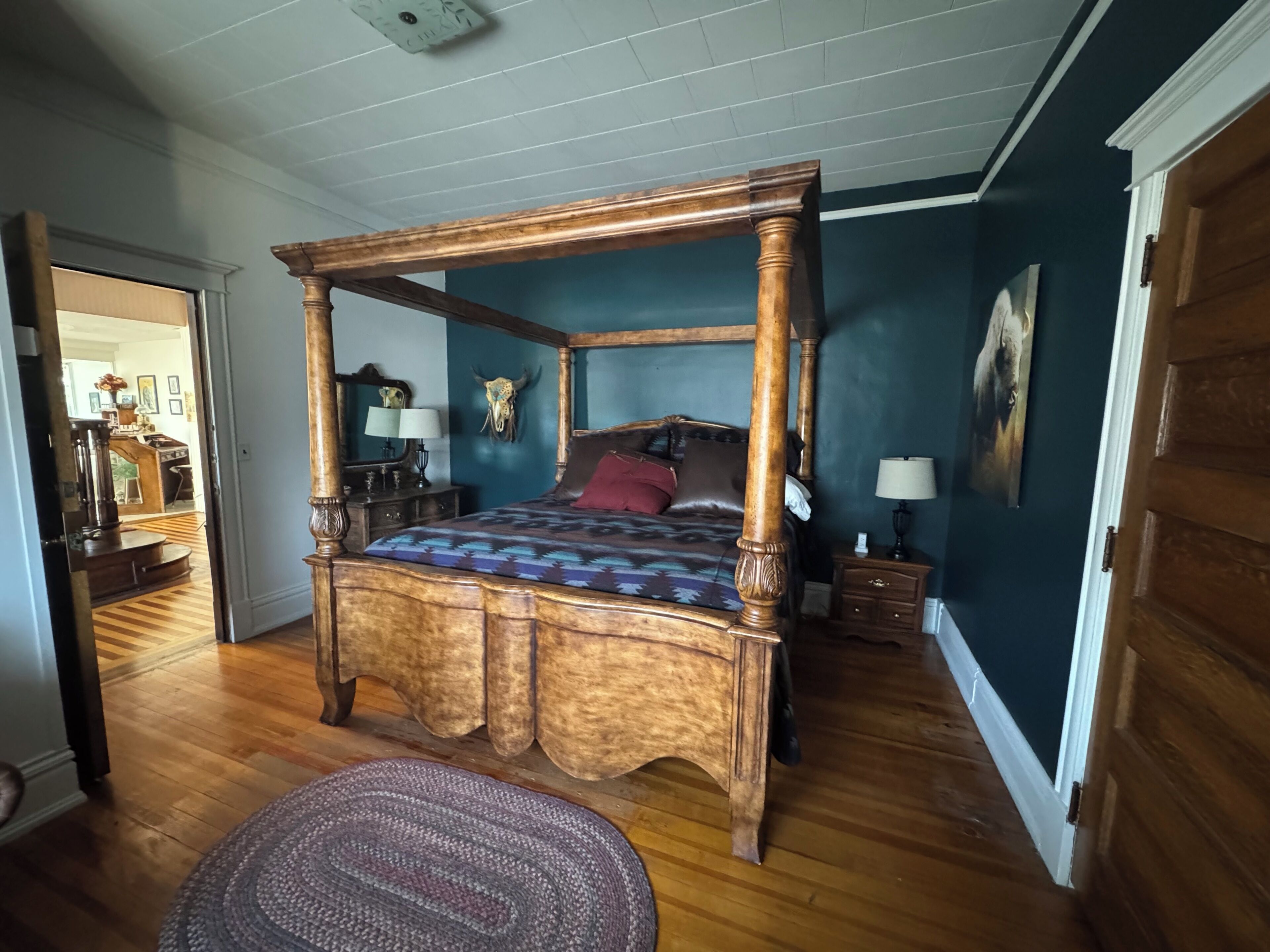 A four-poster bed with a wooden frame, a patterned bedspread, and a red pillow. There is a wooden nightstand with a lamp, a wall-mounted mirror, and a skull decoration. The room has wooden flooring and a door leading to another room.
