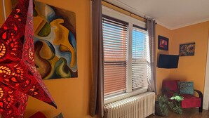 Ein Zimmer mit Heizkörper, einem Fenster mit Jalousien, einem roten Sessel, einem blauen Sessel und einer Wand mit einem Gemälde.