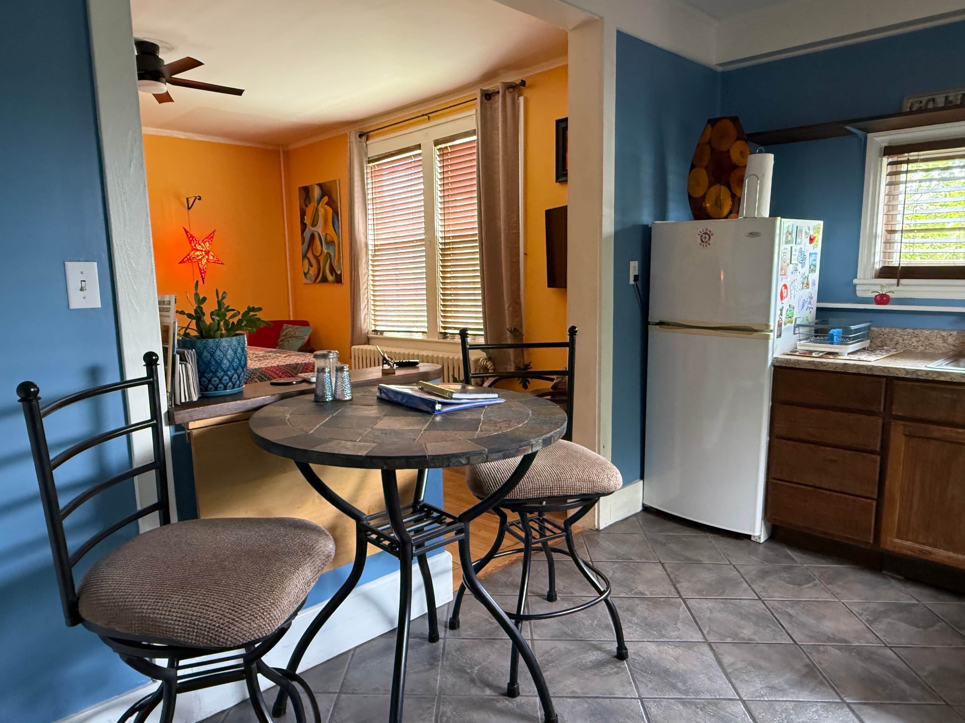 Une cuisine équipée d’une table à manger, de chaises, d’un réfrigérateur et d’une fenêtre avec des stores.