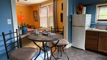 Une cuisine équipée d’une table à manger, de chaises, d’un réfrigérateur et d’une fenêtre avec des stores.