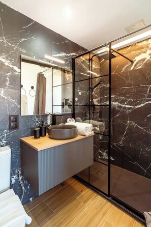 A modern bathroom with a marble wall, a glass shower enclosure, a wooden vanity, and a large mirror.
