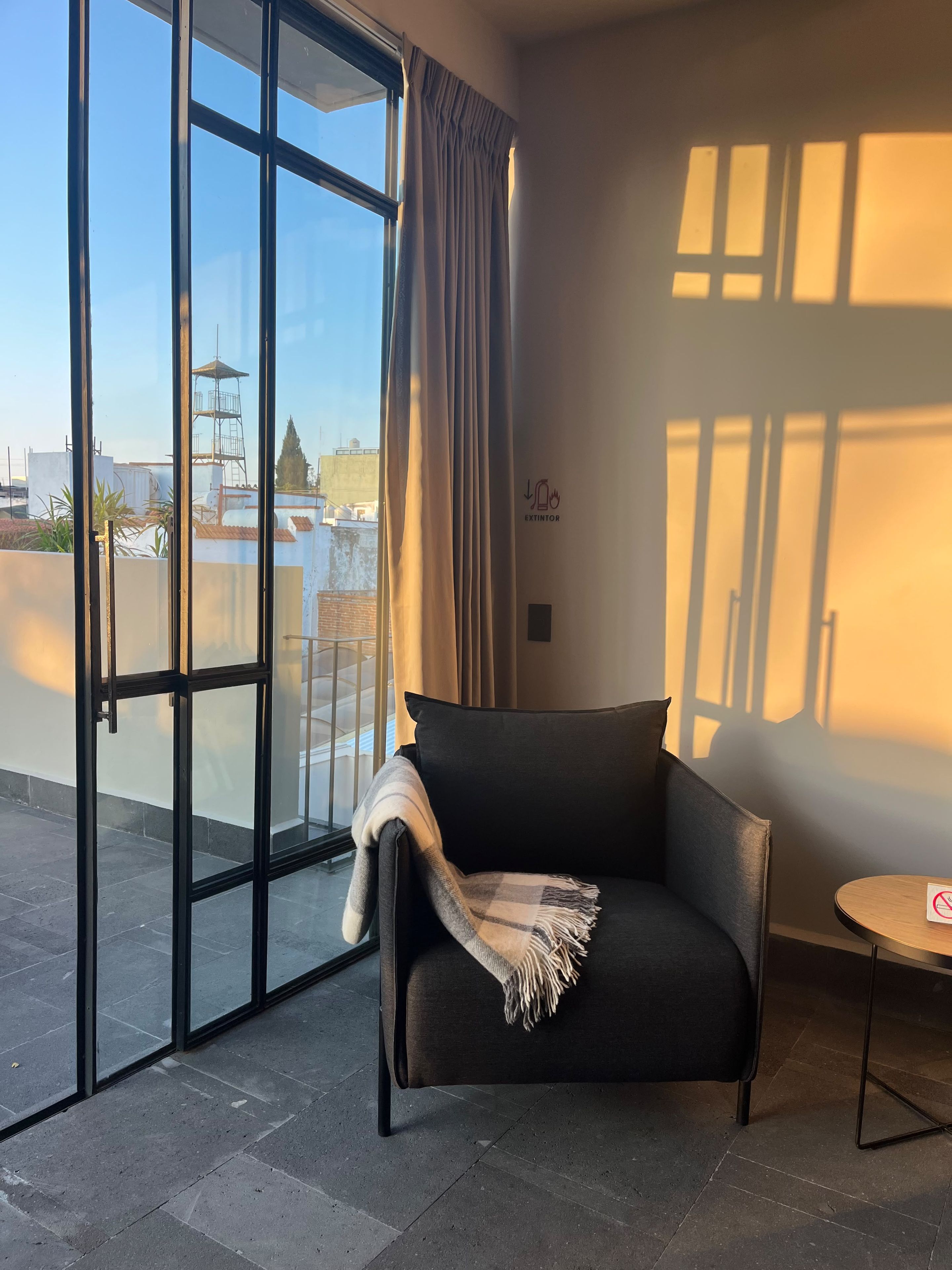 Una habitación moderna con un sofá de cuero negro, una pequeña mesa redonda y un amplio ventanal corredero que da a una terraza exterior.