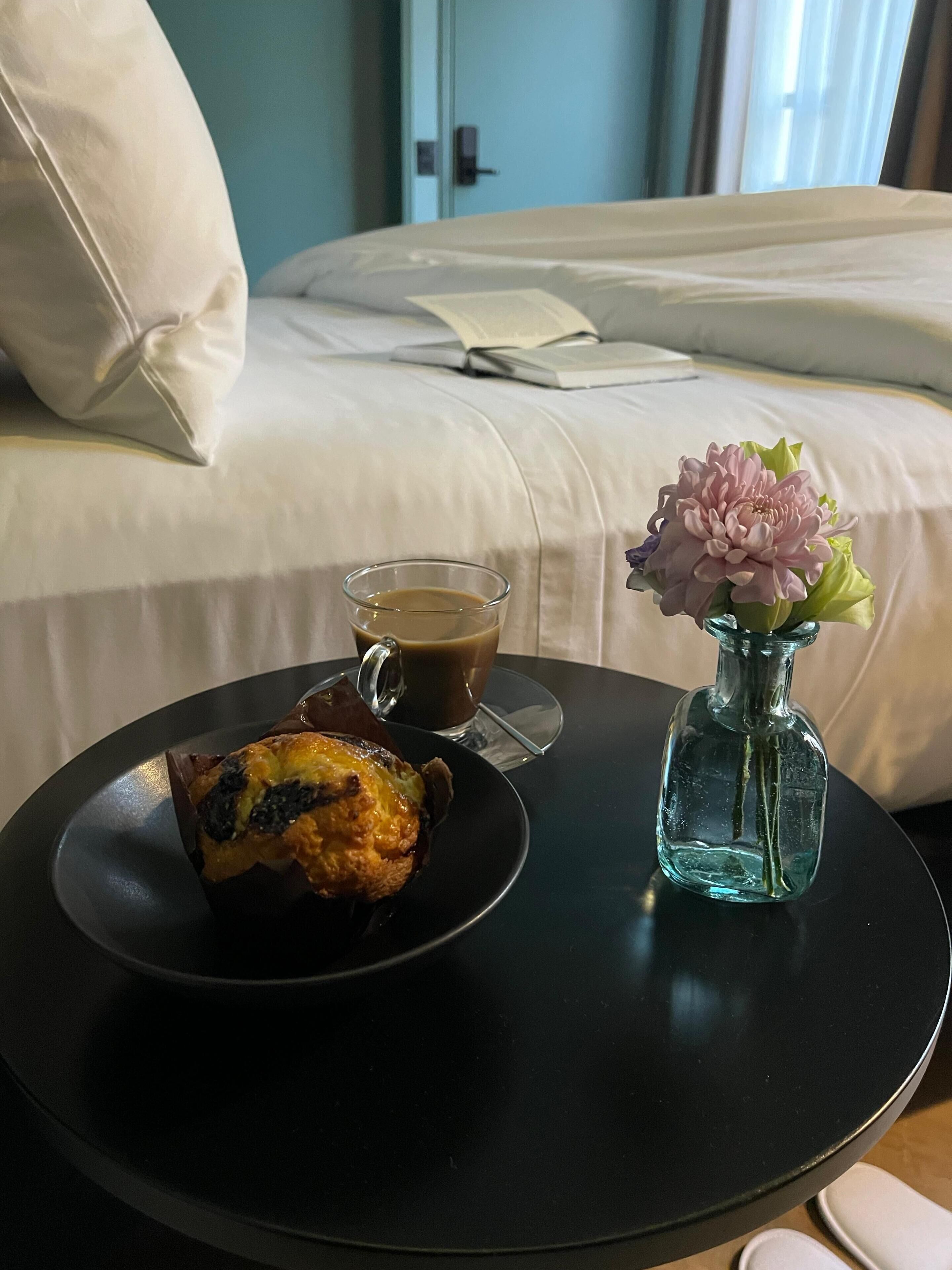 Een bed met witte lakens, een nachtkastje met een glas koffie, een muffin en een vaas met bloemen.