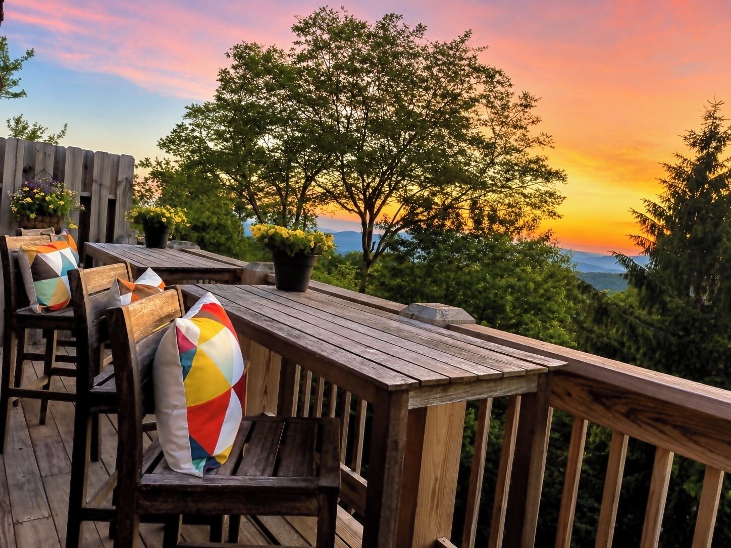 An outdoor deck with wooden furniture and potted plants, overlooking a scenic view.