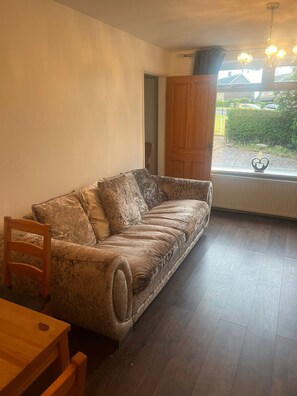 A room with a sofa, a wooden chair, a table, a window with a view of the outside, and a radiator. - Comfortable 3 Bedroom House-ng5,sleeps 5 (Nottingham)