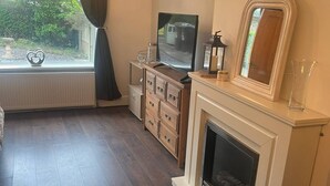 Ett rum med en öppen spis, en tv på ett stativ, en spegel och ett fönster med gardiner.