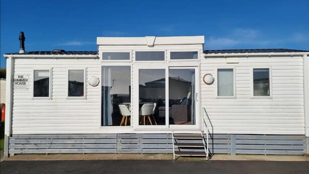 Una casa móvil blanca con un letrero que dice "The Summer House". Tiene una puerta principal, dos ventanas y una pequeña zona de porche con un banco.
