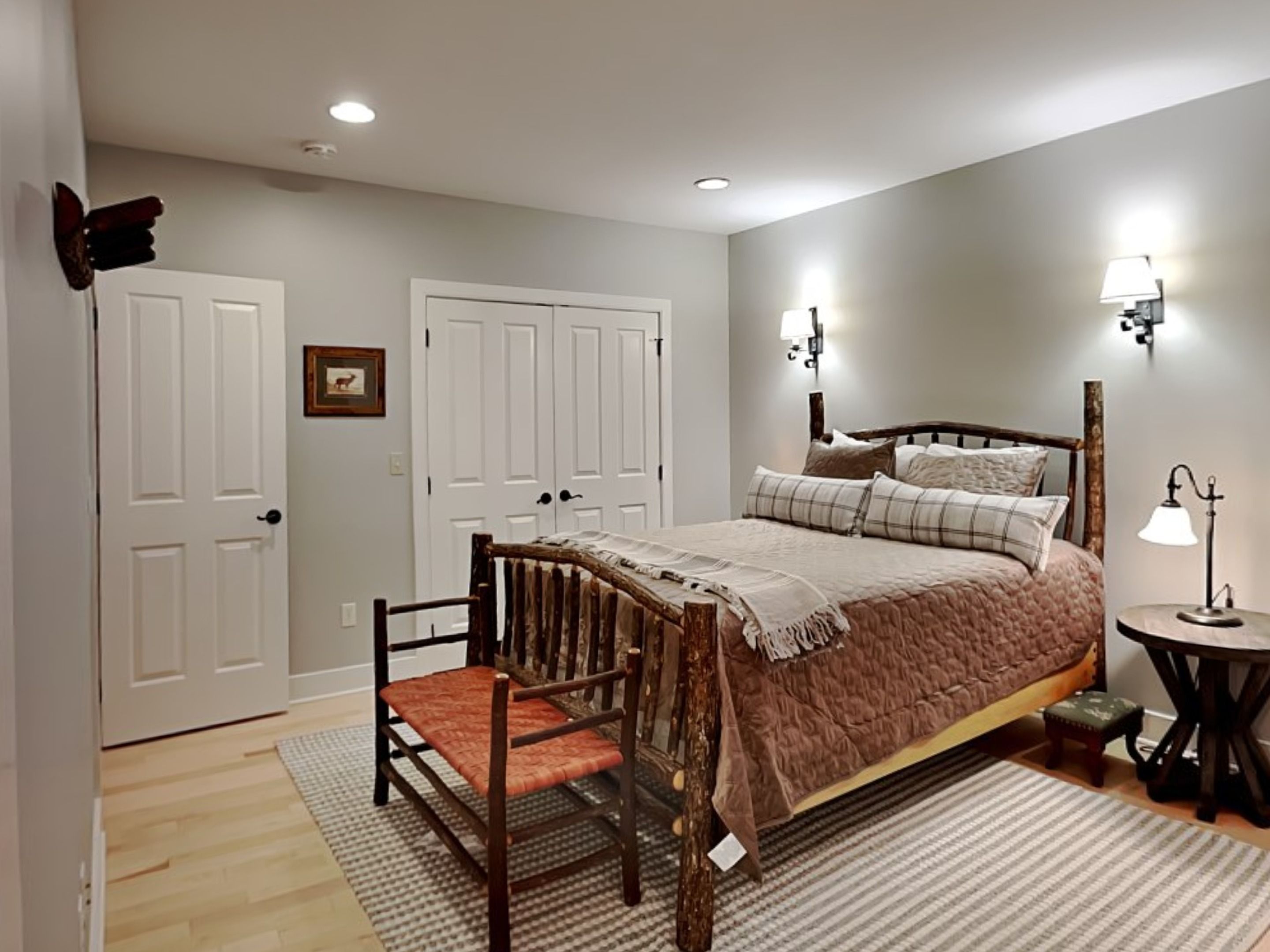 A bedroom with a wooden bed, two white doors, a small table with a lamp, and a chair.